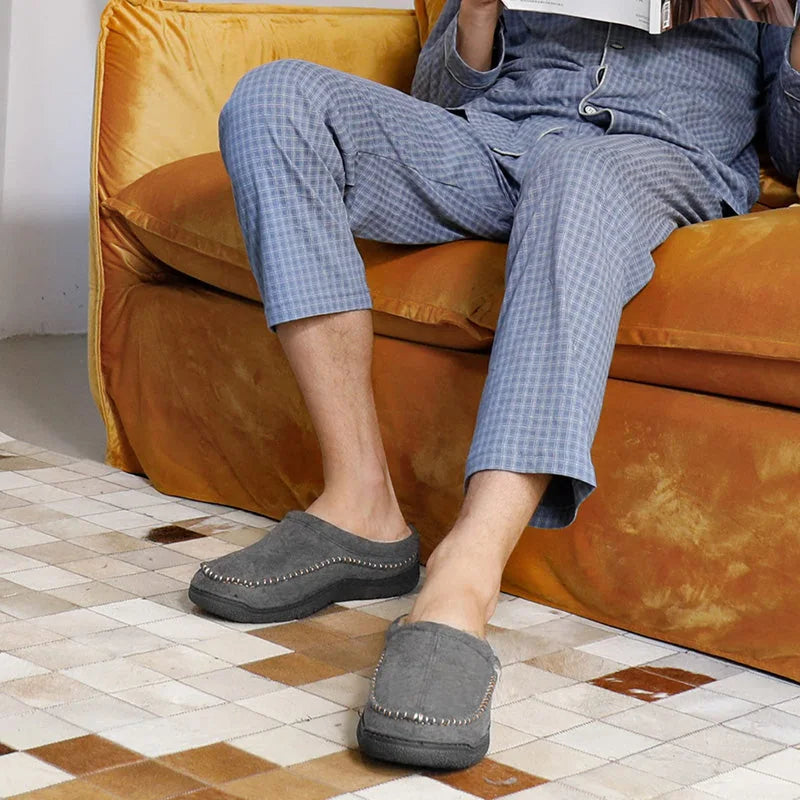 Grey wide fit house slippers worn with blue pyjamas beside a sofa