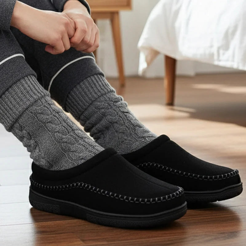 All-black house slippers worn with grey socks beside a bed