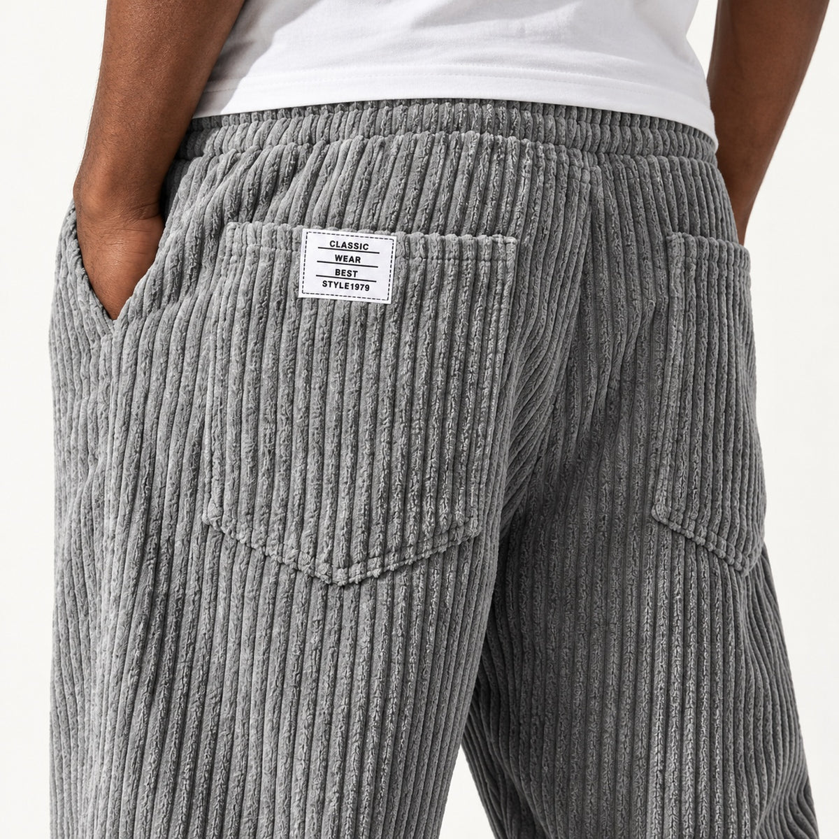 Close view of the grey trousers showing the vertical rib texture, elasticated waist and rear patch pockets, with a white sewn label on the wearer’s left rear pocket