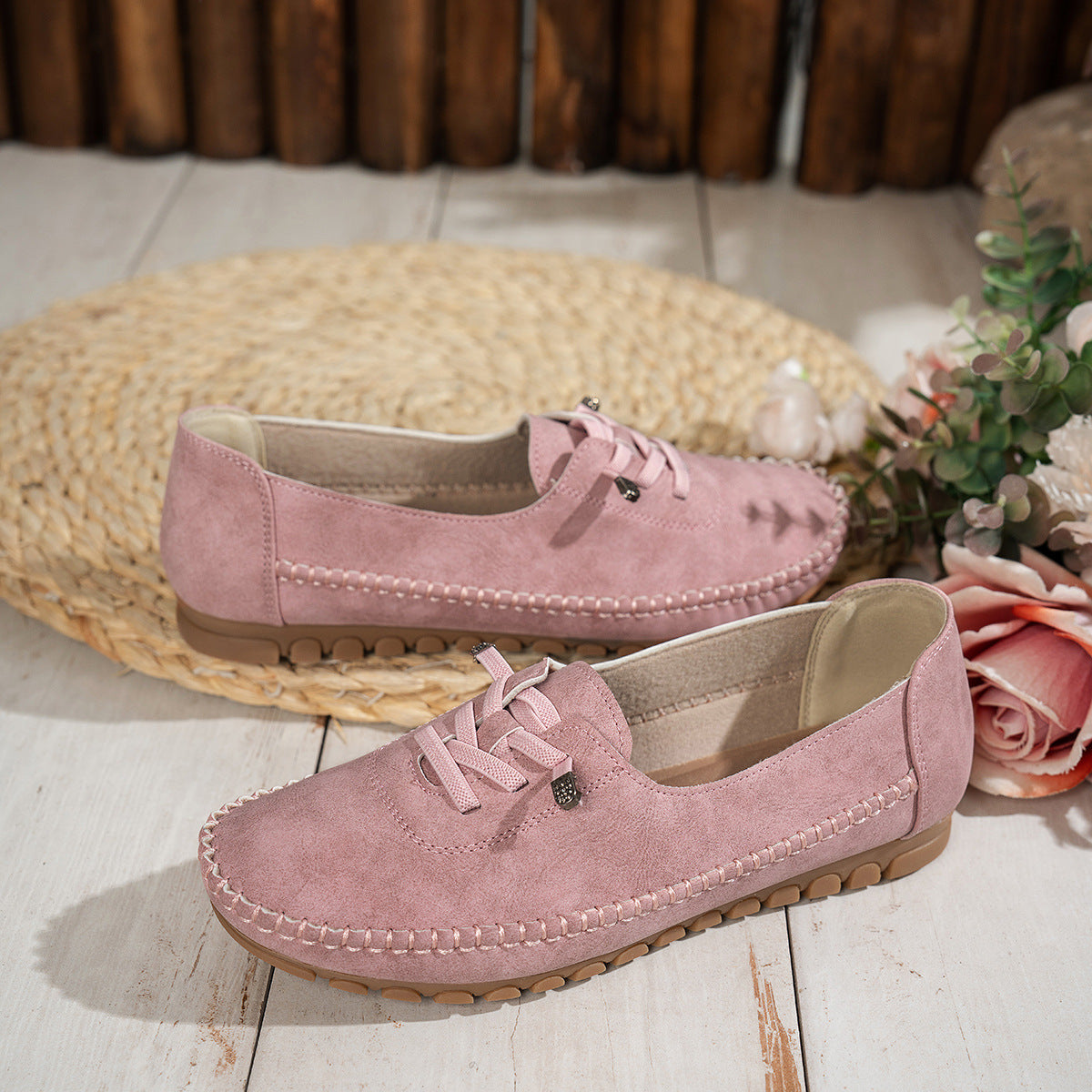 Pink women's suede loafers with decorative lacing, displayed beside flowers