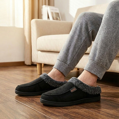 Auldweave Men’s warm wide fit winter slippers. Person wearing black slippers with grey fur trim in a living room setting.