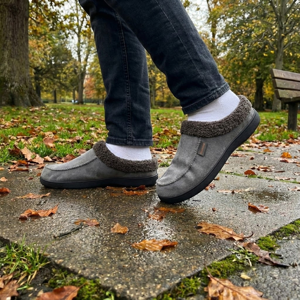 Auldweave Men’s warm wide fit winter slippers. Person wearing grey slippers with fur trim on a sidewalk with autumn leaves.