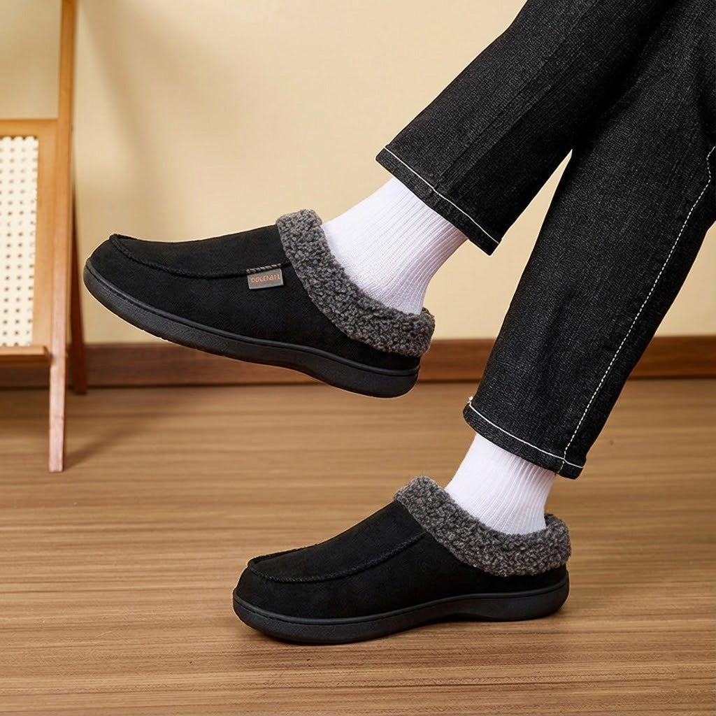 Auldweave Men’s warm wide fit winter slippers. Person wearing black slippers with grey fur trim on a wooden floor.