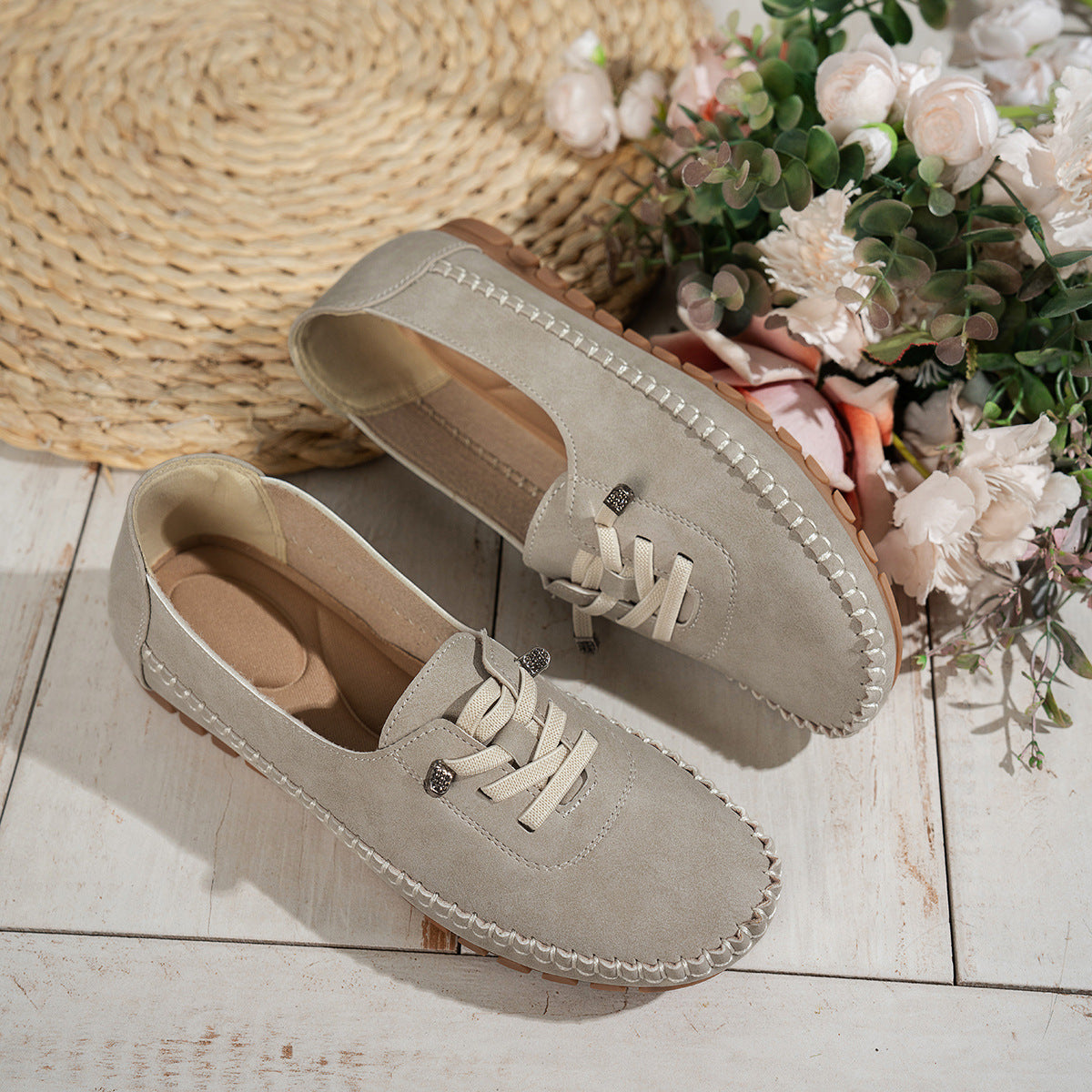 Light grey women's suede loafers with stitched edges, displayed beside flowers
