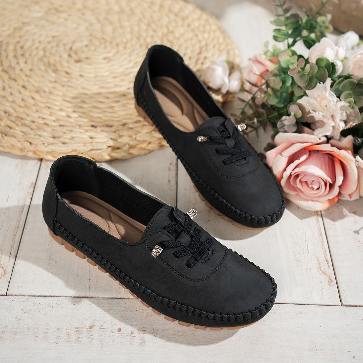 Black women's suede loafers with decorative lacing, displayed beside flowers