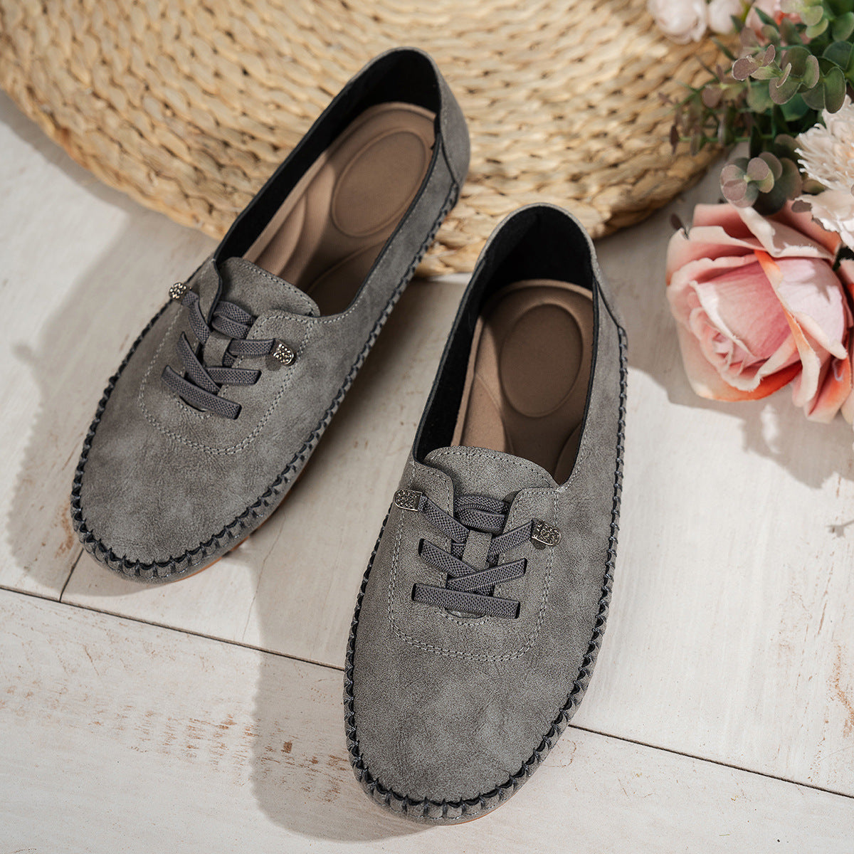 Dark grey women's suede loafers with decorative lacing, top view beside flowers